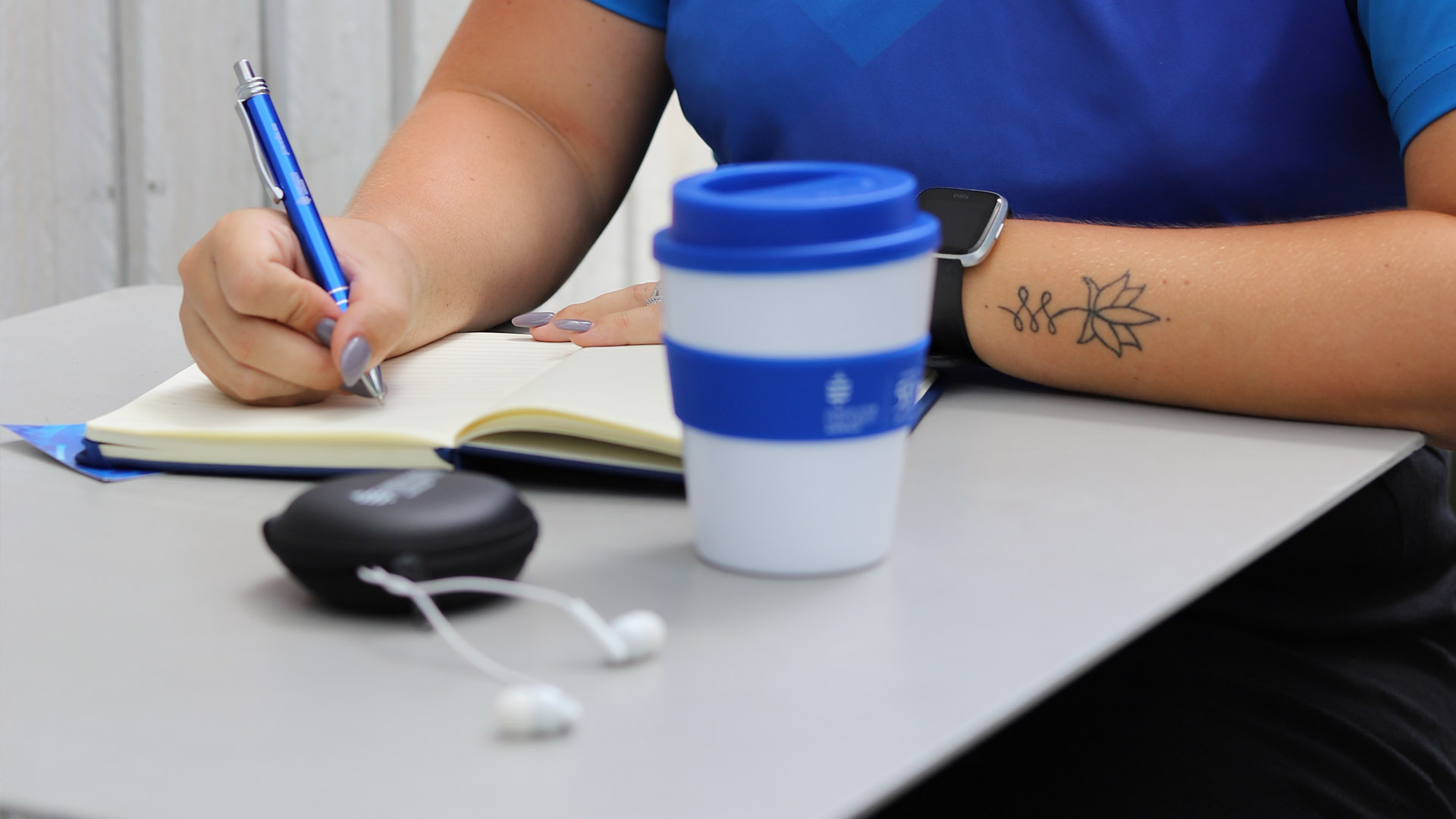 A James Cook University branded reusable coffee cup sits on a cafe table next to a notebook. A JCU student's hand is visible writing in the notebook. 