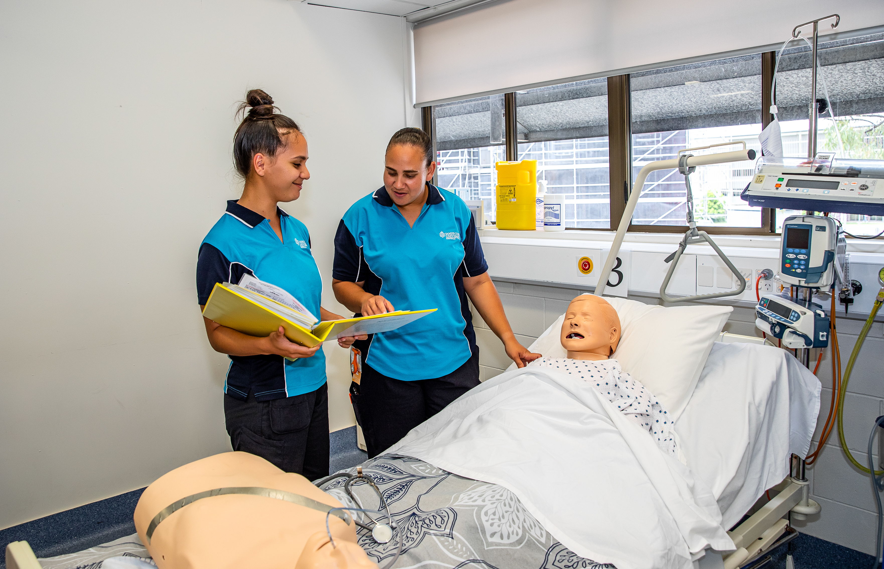 Two female James Cook University Bachelor of Nursing Science students stand over a hospital bed examining a medical model. 