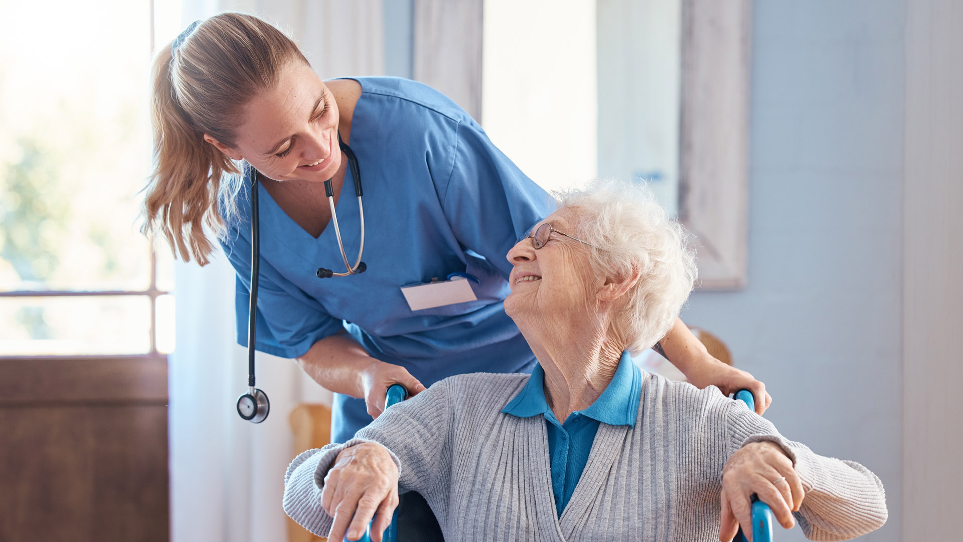 A nurse leans over to speak to her elderly female patient in a wheelchair. 