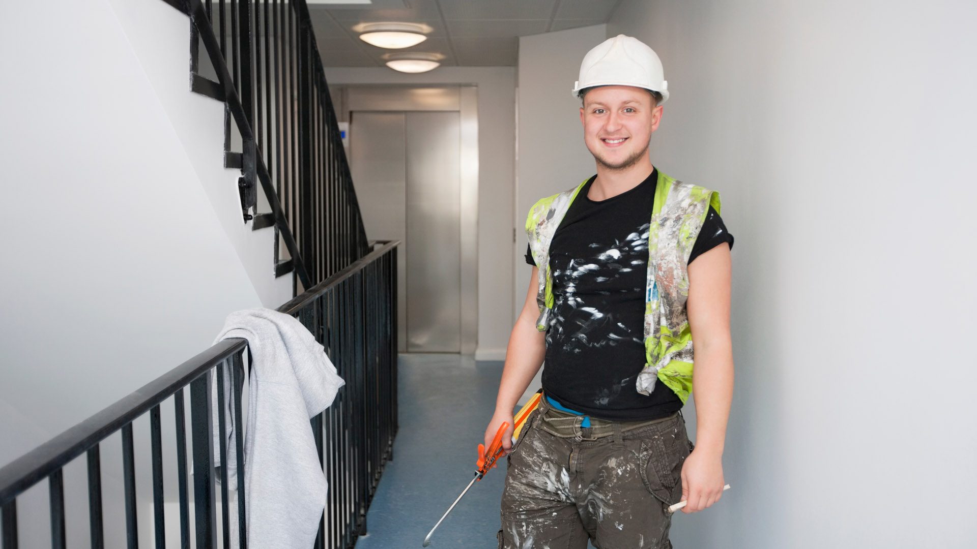 A young tradesperson stands holding a painting tool, wearing a hard hat and high-visibility vest splattered with paint. 