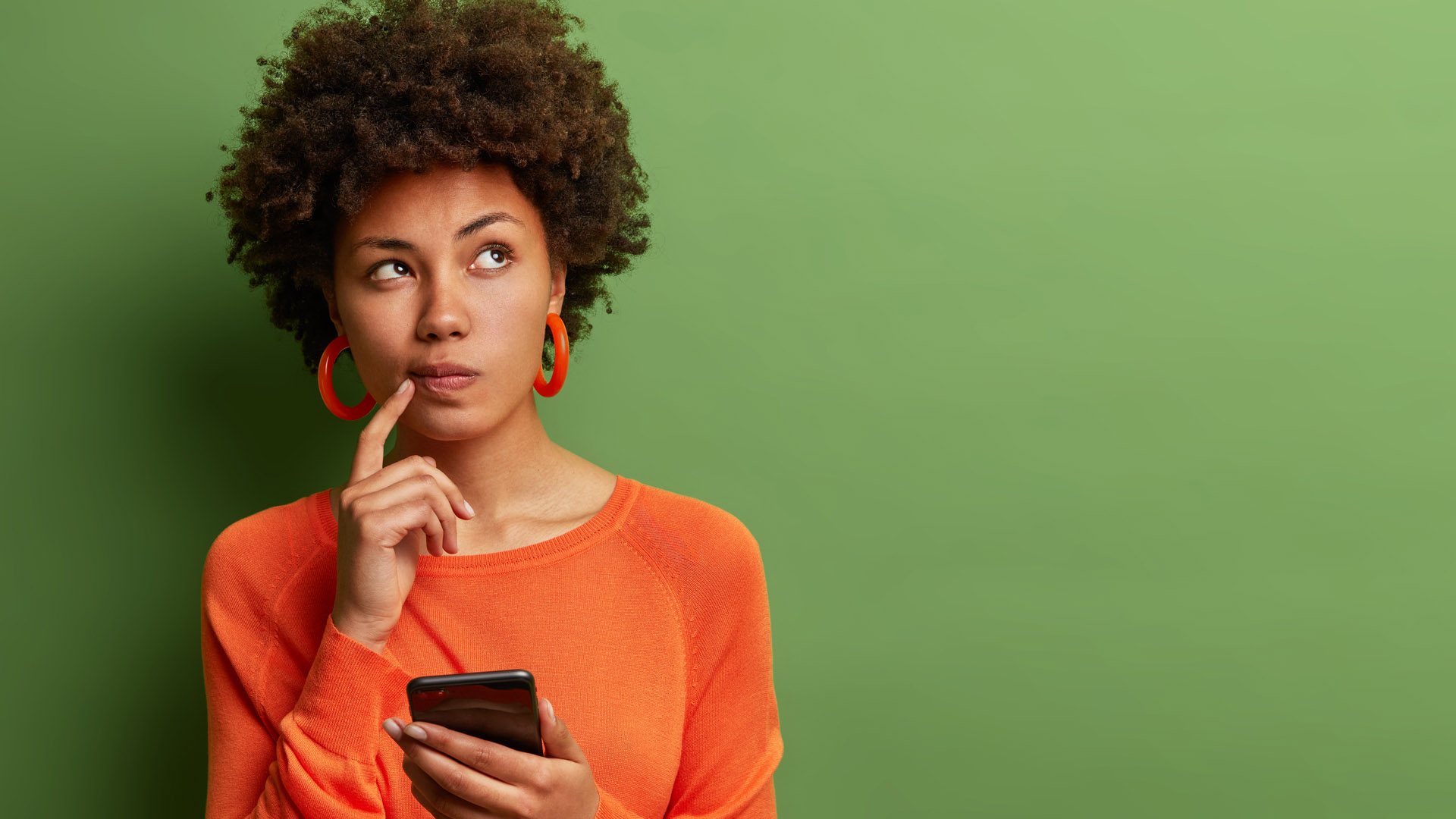 A woman in an orange shirt stands with one finger on her mouth, holding a phone and looking like she wants the answer to a question. 