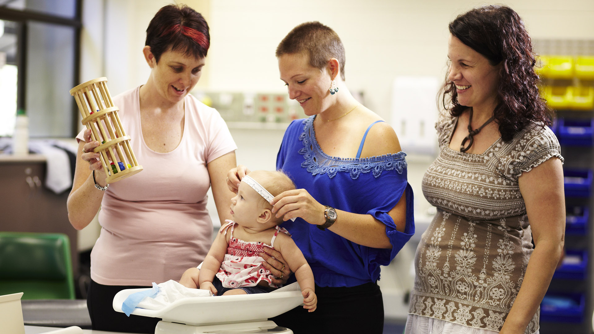 A midwife and mothers hold an infant on an examination table to be measured. 